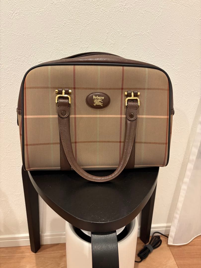 Front view of the brown Burberrys Nova Check canvas handbag with leather handles and gold hardware placed on a small stool against a wall.