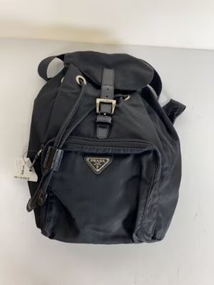 Front view of a black PRADA nylon backpack with flap closure, front zip pocket, and triangular logo.